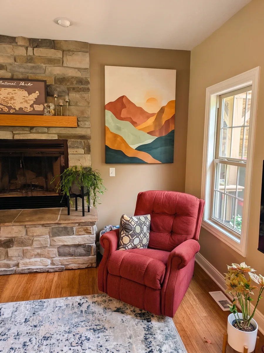 A cozy living room corner with a warm mountain canvas above a red armchair beside a stone fireplace